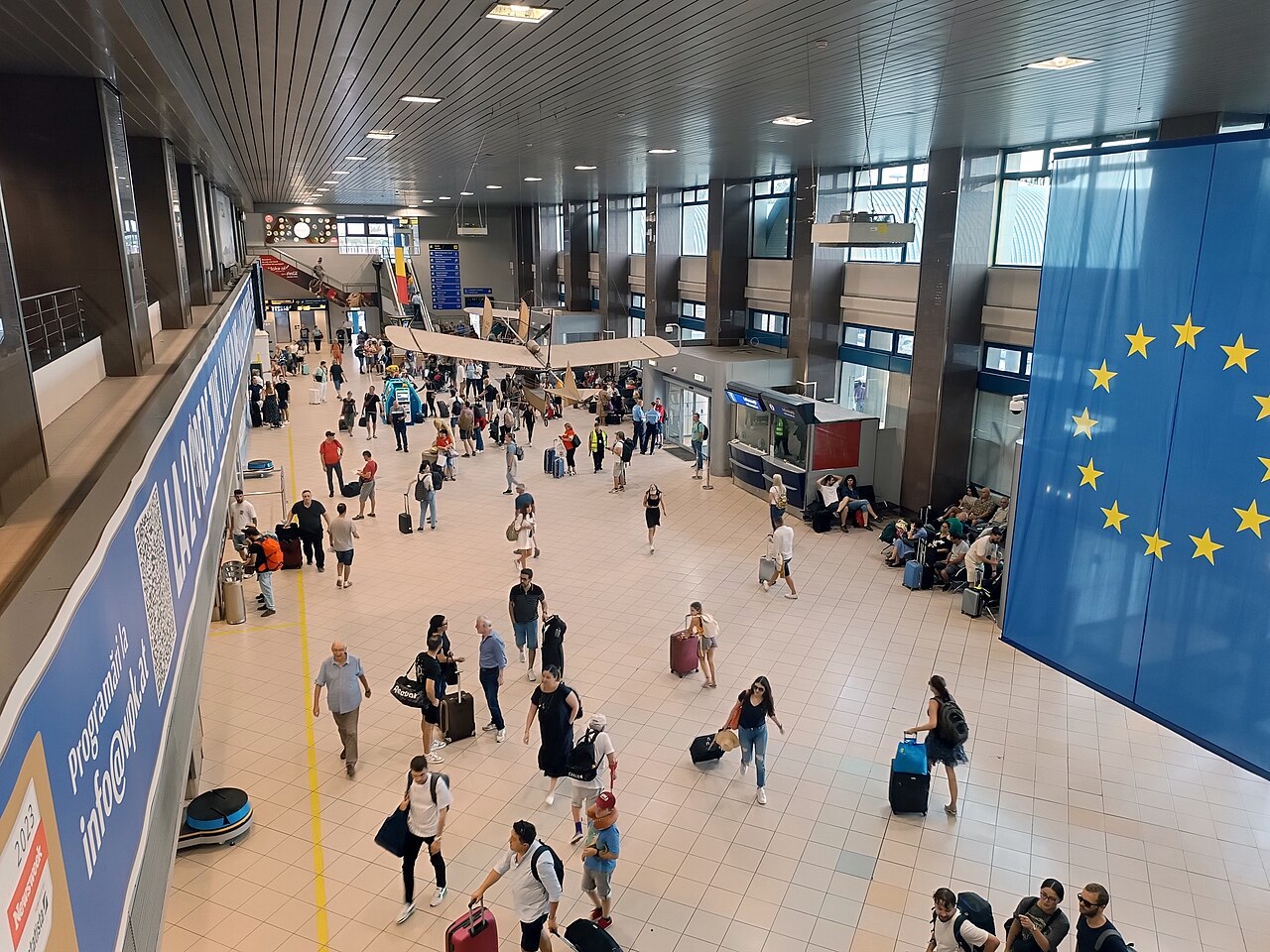 Hala letiskového terminálu s cestujúcimi, batožinou a veľkou vlajkou Európskej únie