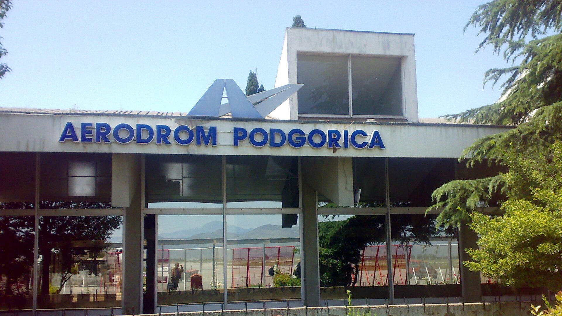 Vchod do terminálu s modrým nápisom Aerodrom Podgorica nad presklenou fasádou